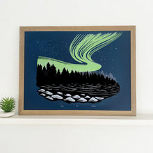 Load image into Gallery viewer,  Northern lights artwork. A rich neon green aurora ink on top of a dark blue backround behind the shadows of a Black Forest. A lake in the foreground showing some light on rocks. Framed photo of aurora borealis
